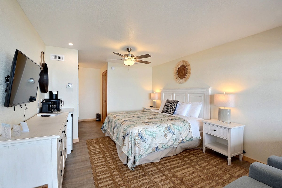 A hotel room with a bed, a TV, a desk with a coffee maker, and a ceiling fan.