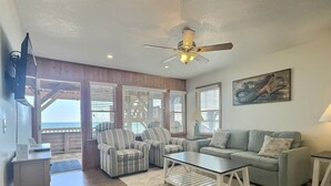 A living room with a sofa, armchairs, a coffee table, and a ceiling fan. There are large windows with blinds, a painting on the wall, and a wooden floor. - Lighthouse View Oceanfront Lodging (Buxton)