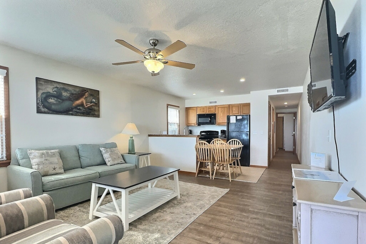 A living room with a sofa, coffee table, and a dining area with chairs. There is a kitchen in the background with a microwave and refrigerator.