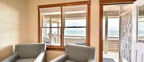 A room with two grey armchairs, a wooden-framed window with a view of the beach, and a door leading to a wooden deck.