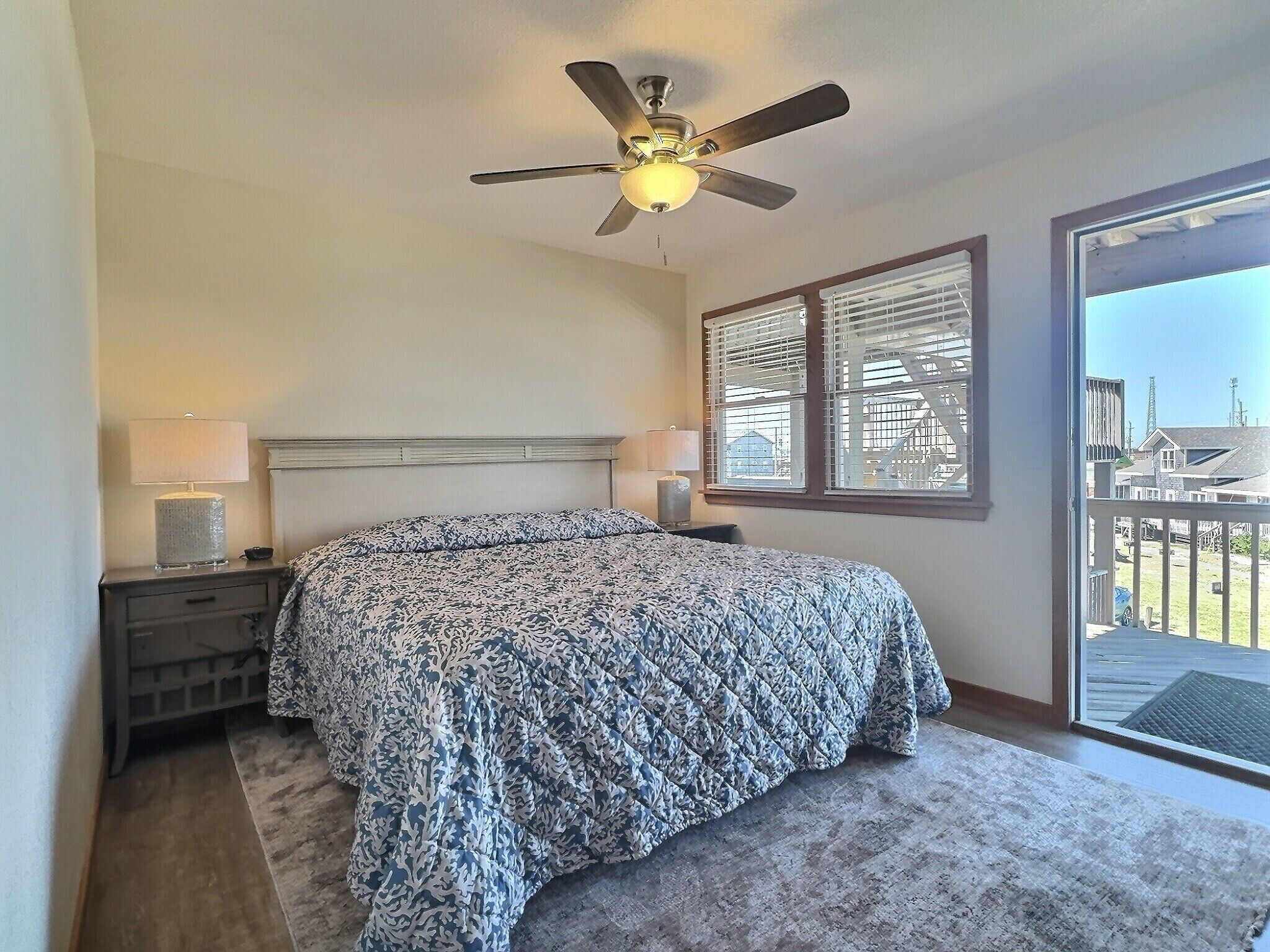 A bedroom with a bed, two bedside lamps, a ceiling fan, and a large window with blinds.