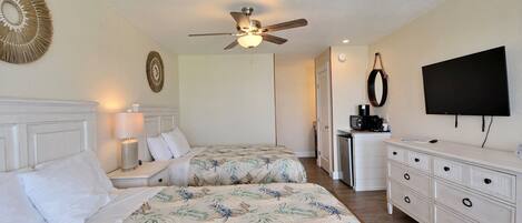 A hotel room with two beds, a ceiling fan, a TV, a dresser, and a desk with a lamp.