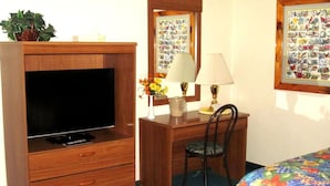 A room with a wooden TV stand, a desk with two lamps, a chair, and two beds with floral bedding. - Color Country Motel (Panguitch)