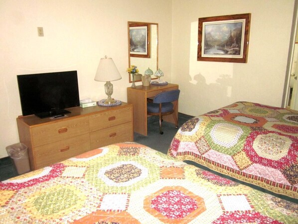 A hotel room with a TV, a desk with a chair, two beds with quilts, and two framed pictures on the wall.