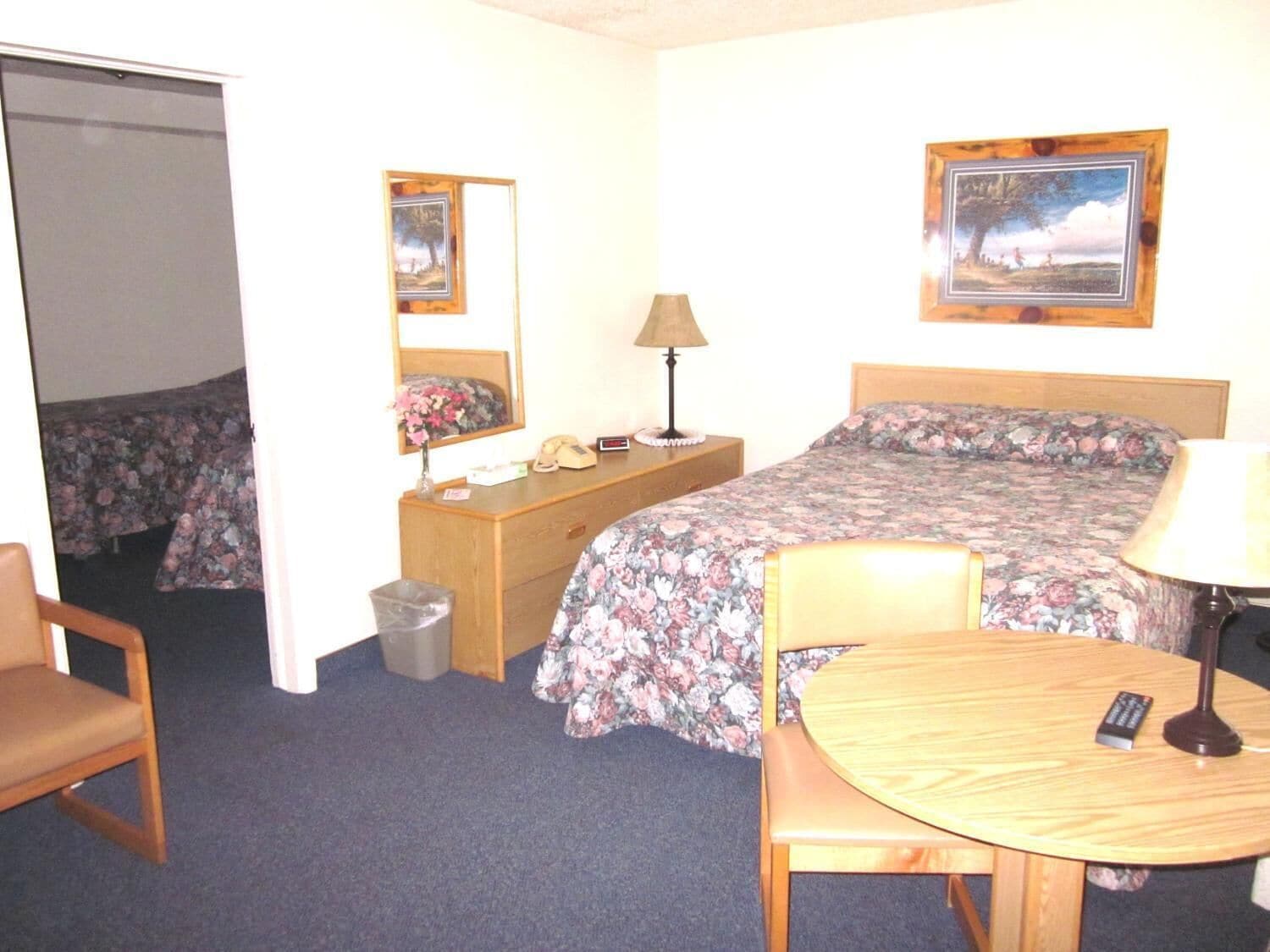 A hotel room with a bed, a desk with a lamp, a chair, a small table, and a trash bin.
