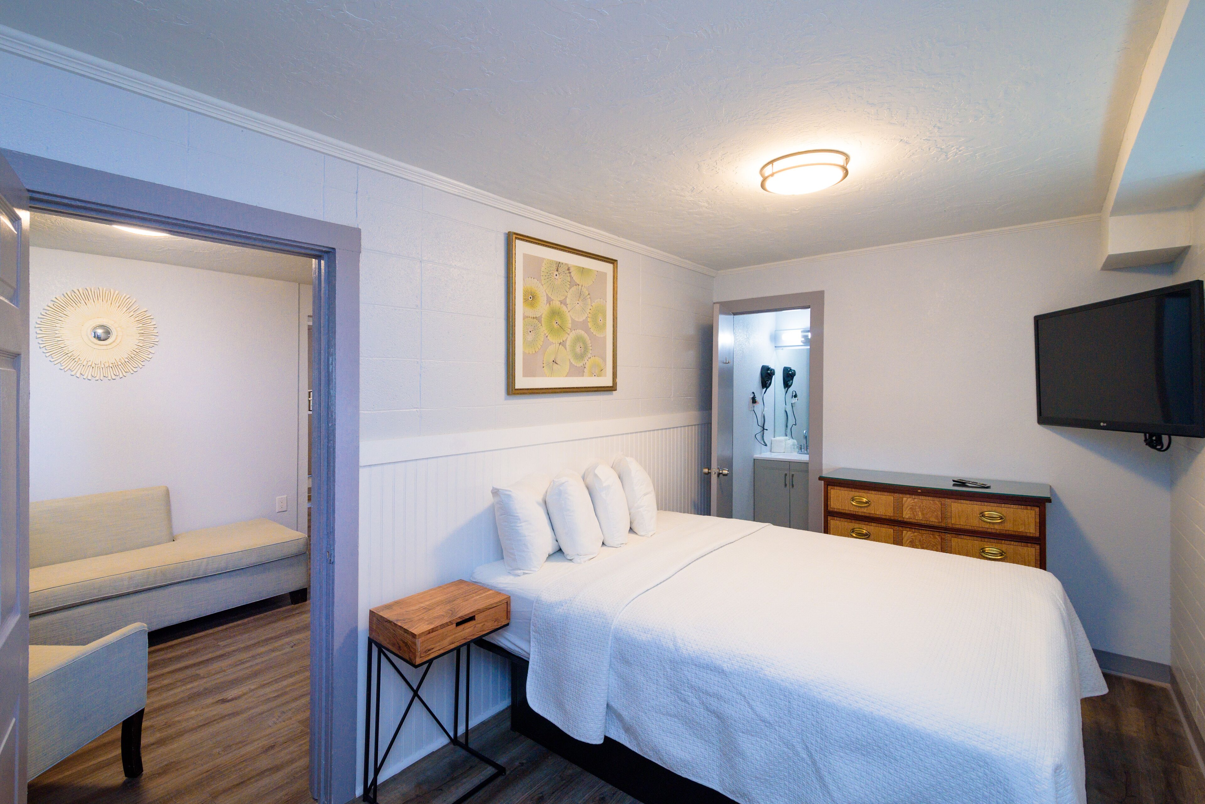 A bedroom with a bed, a wooden nightstand, a dresser, a television, and a door leading to another room.