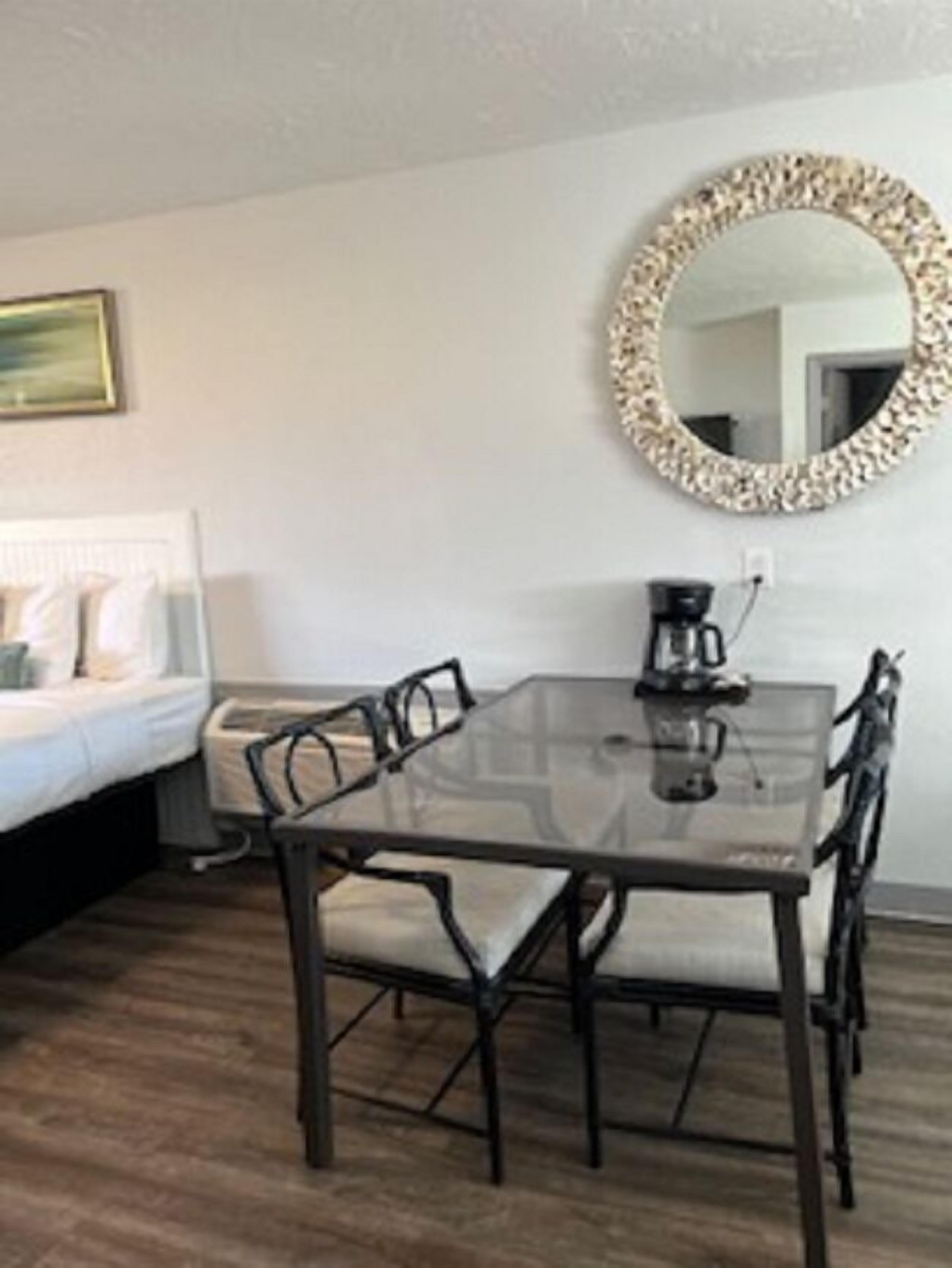 Una habitación de hotel moderna con una mesa de comedor de vidrio, cuatro sillas, una cafetera, un espejo redondo y una cama con ropa de cama blanca.