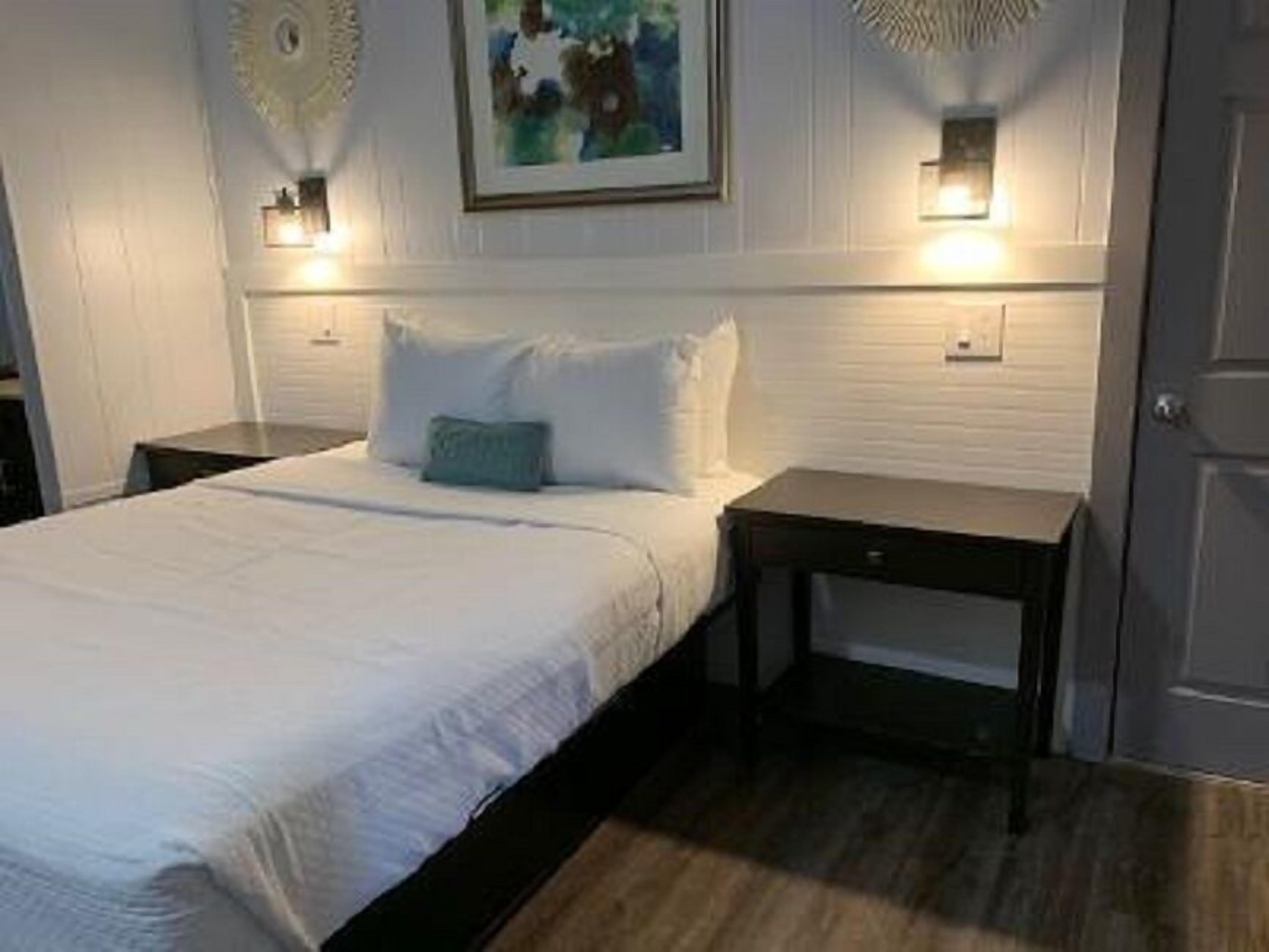 Uma cama bem arrumada com roupa de cama branca e um travesseiro azul-turquesa, com duas mesas de cabeceira e luminárias fixadas na parede.