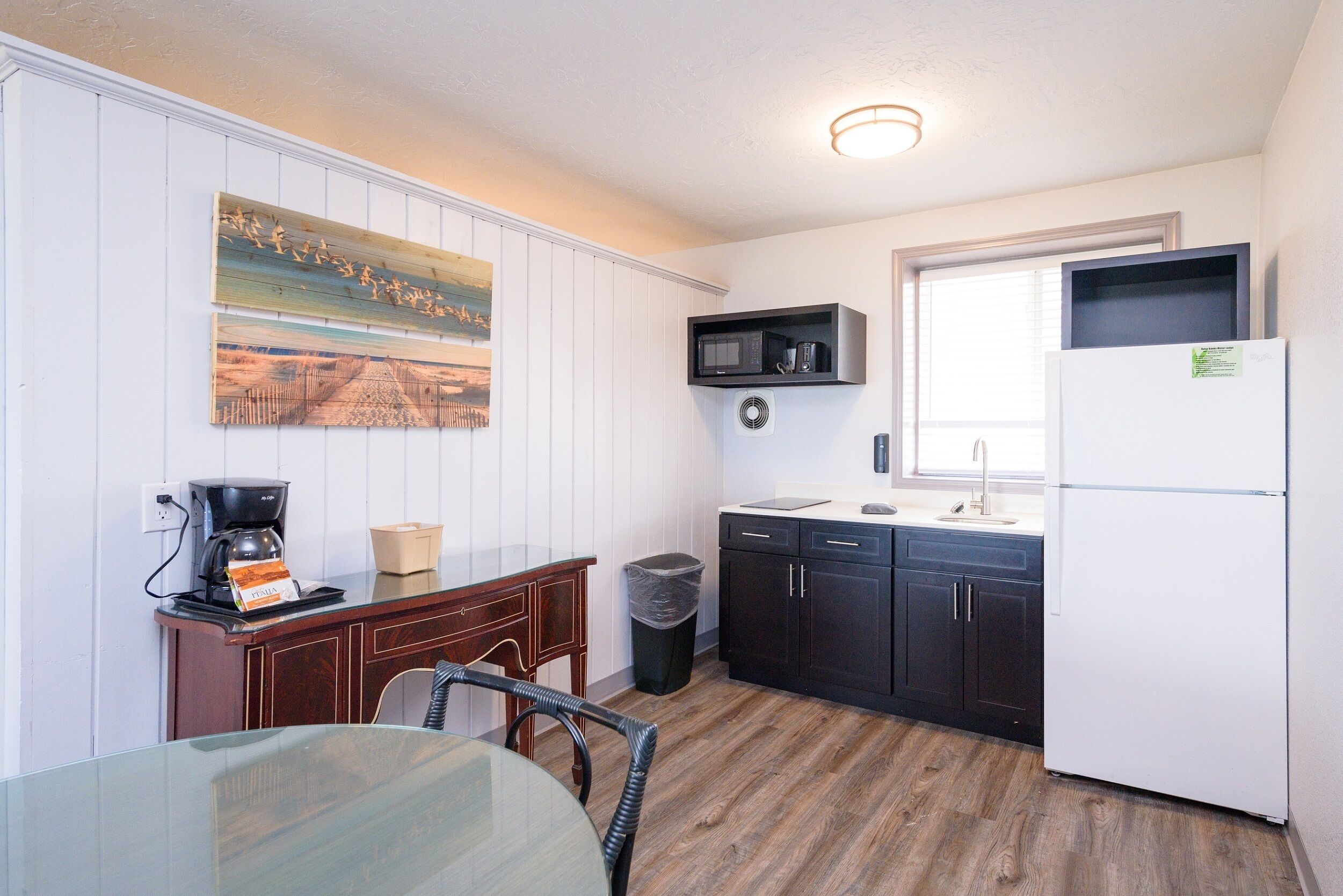 A compact kitchen with wooden flooring, a dining table, a coffee maker, and a microwave on a shelf.