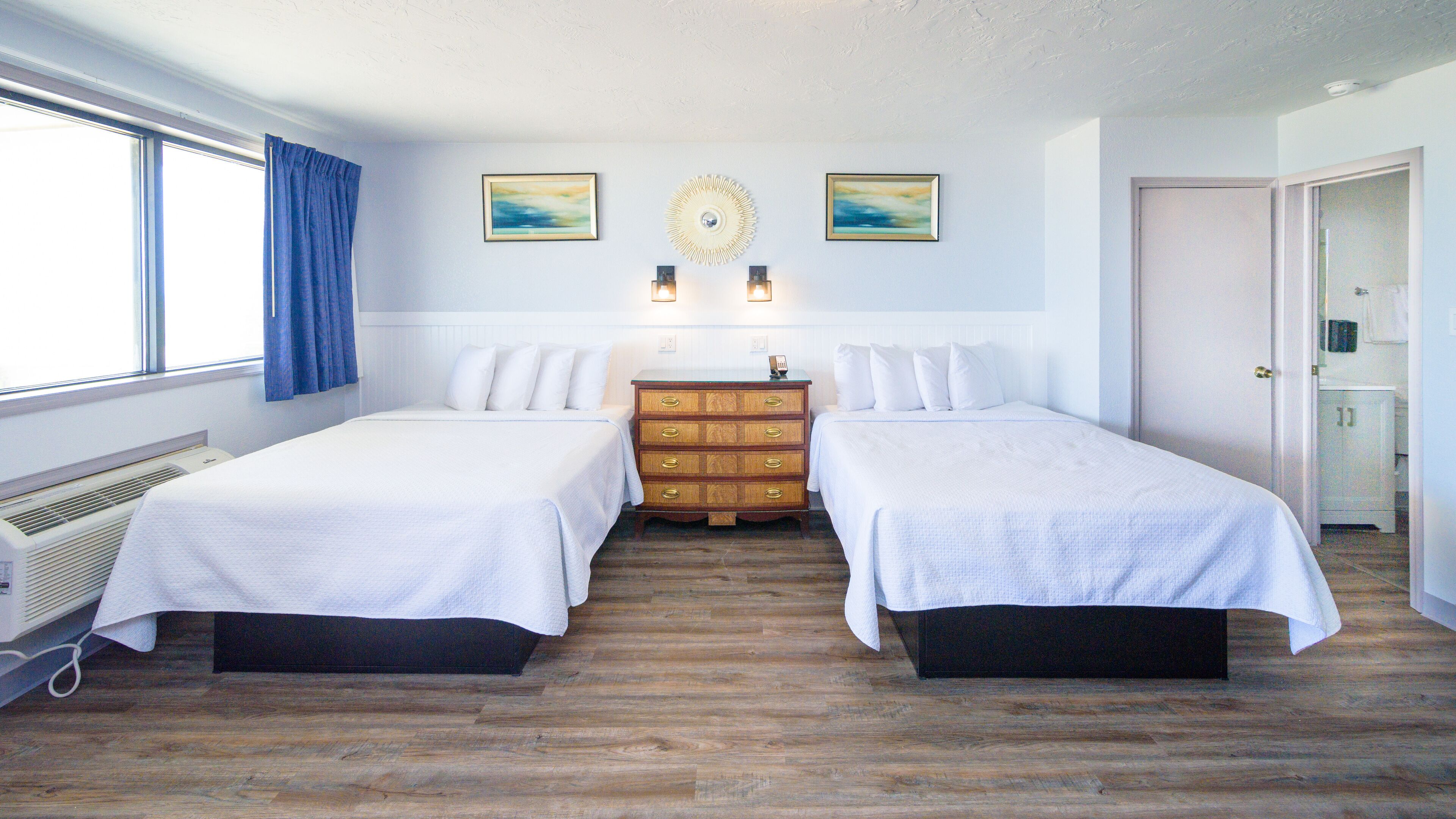 A hotel room with two single beds, a wooden dresser, a radiator, and a door leading to a bathroom.