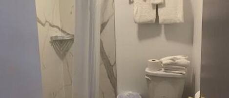 A bathroom with a toilet, a shower curtain, a towel rack with towels, and a trash bin.
