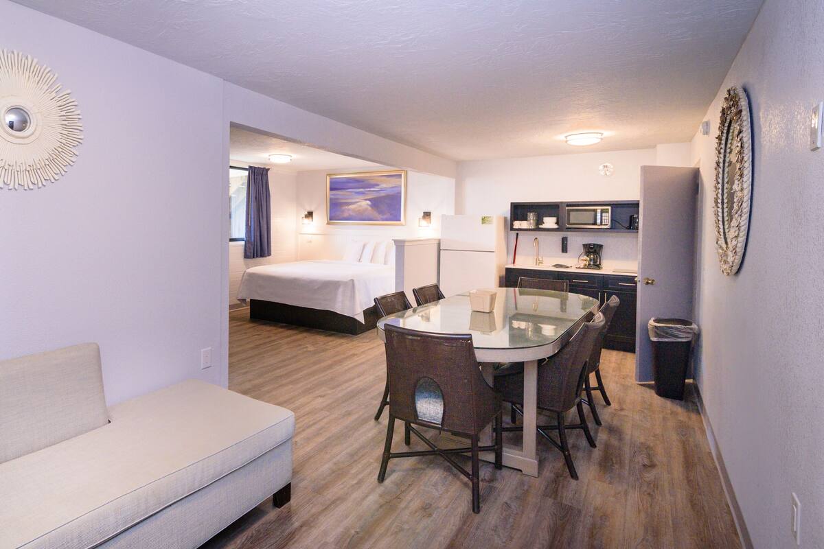 A compact hotel room with a bed, a dining table with chairs, a kitchenette, and a sofa.