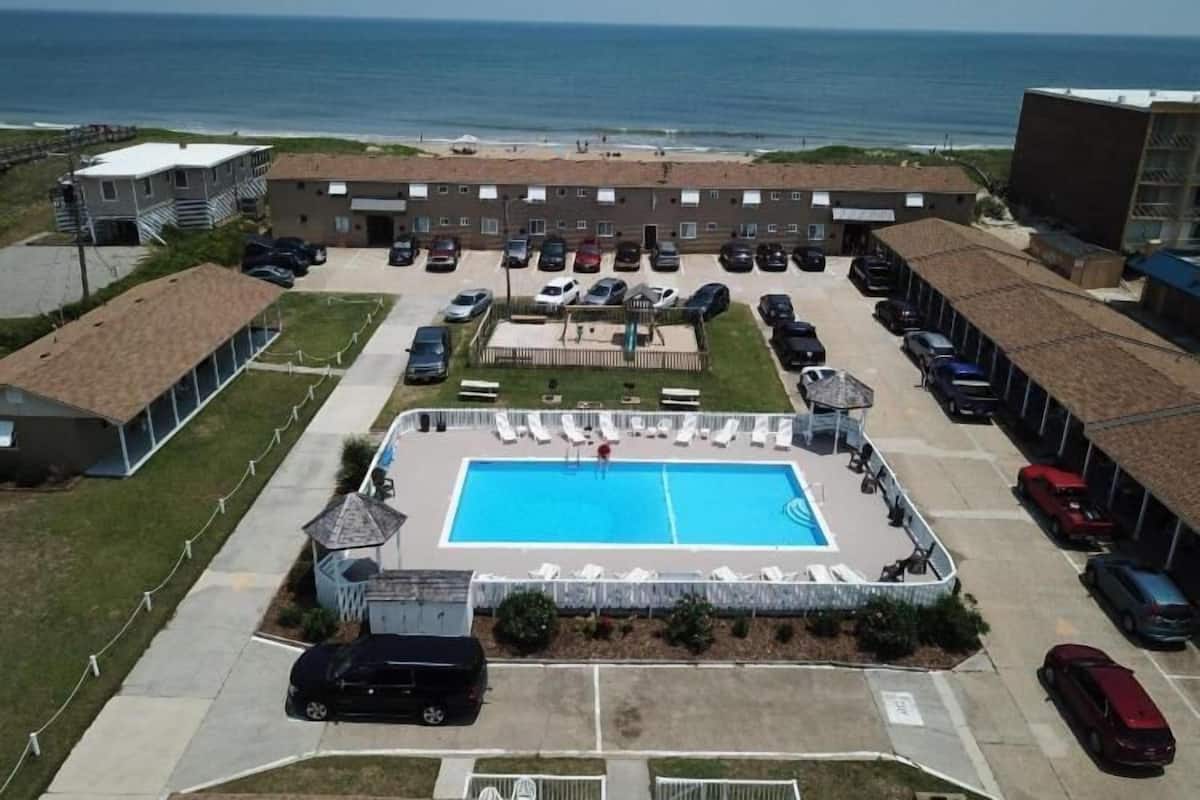 An aerial view of a residential area with a swimming pool, multiple cars, and a beach in the background.