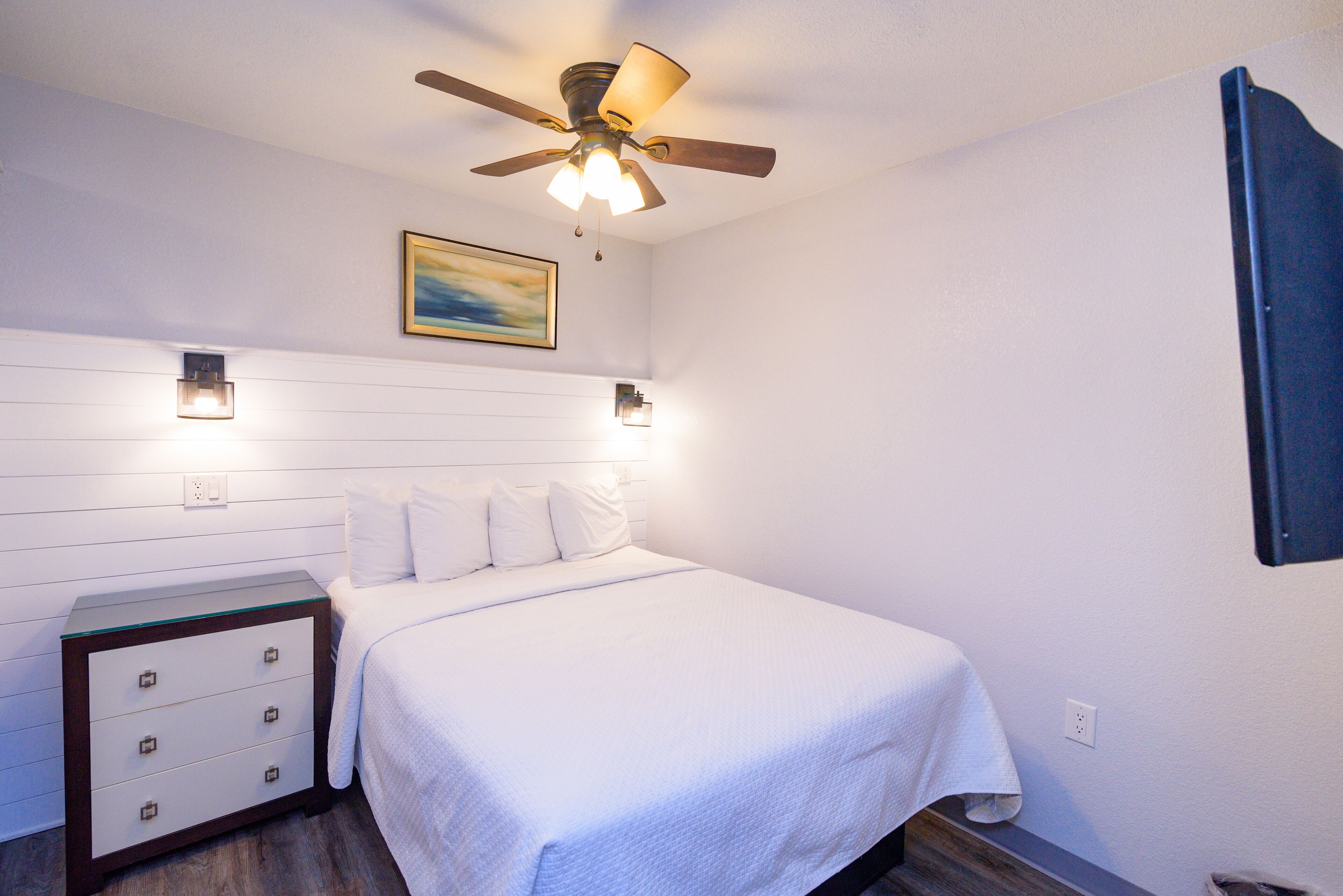 Un dormitorio con cama, mesita de noche, ventilador de techo e iluminación en la pared.