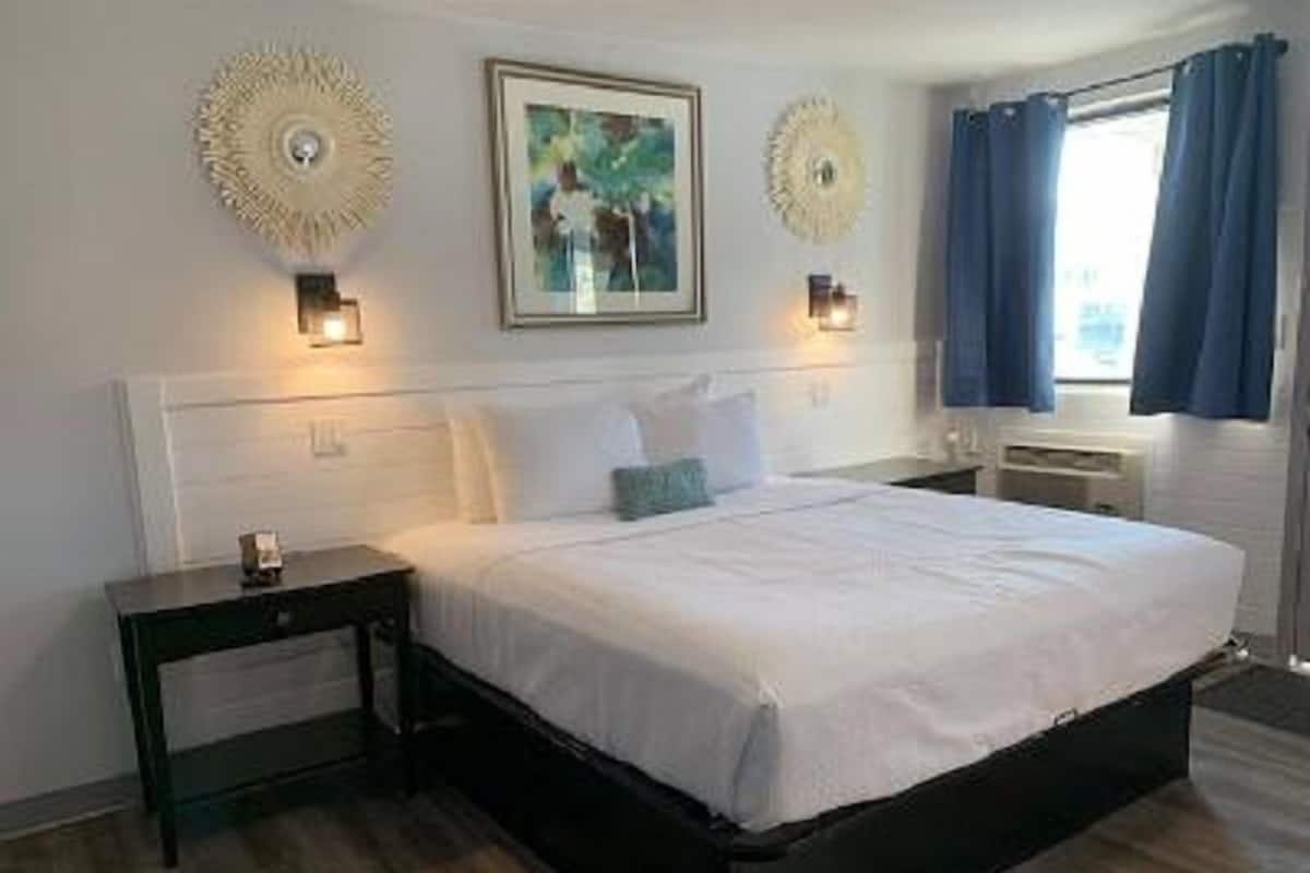 A bedroom with a large bed, two bedside tables, a wall-mounted light fixture, and a window with curtains.