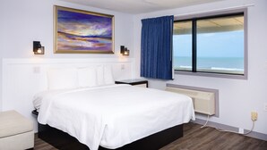 A bedroom with a bed, a nightstand, a bedside table, a window with a view of the ocean, and a painting on the wall. - Outer Banks Motor Lodge (Kill Devil Hills)