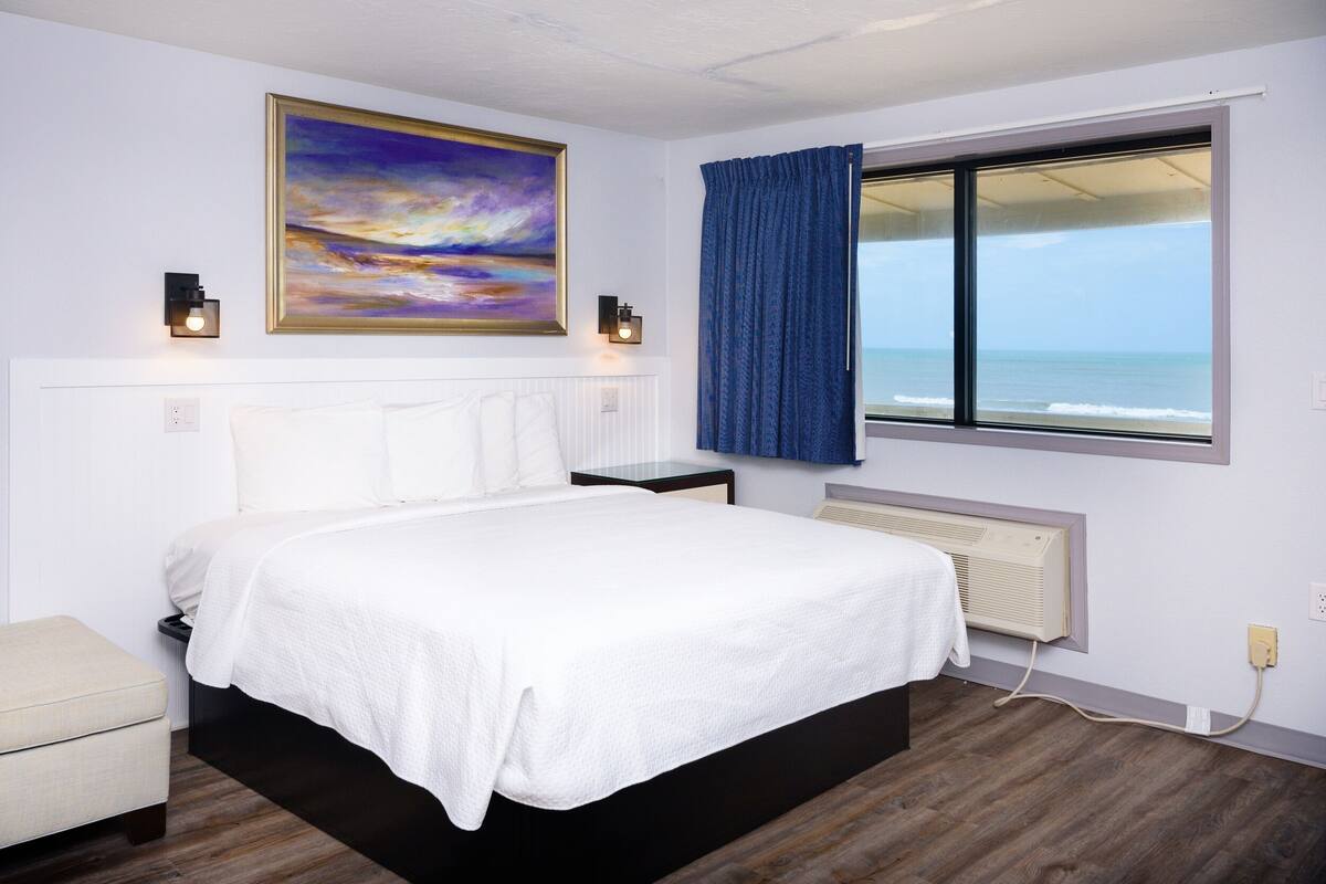 A bedroom with a bed, a nightstand, a bedside table, a window with a view of the ocean, and a painting on the wall.