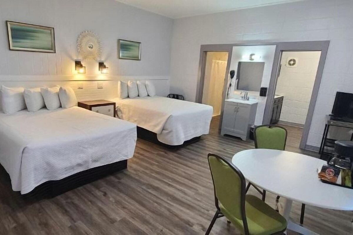 Quarto de hotel com duas camas, uma mesa de jantar, uma televisão e banheiro com espelho e pia.