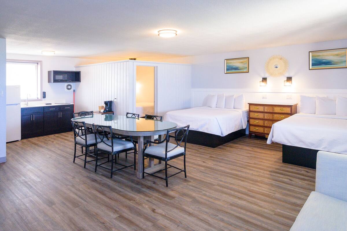 A hotel room with a kitchen area, a dining table with chairs, two beds, and a dresser.