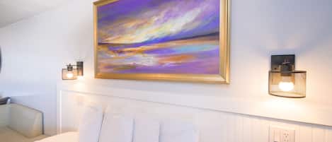 A hotel room with a bed, white bedding, a painting on the wall, and two wall-mounted light fixtures.