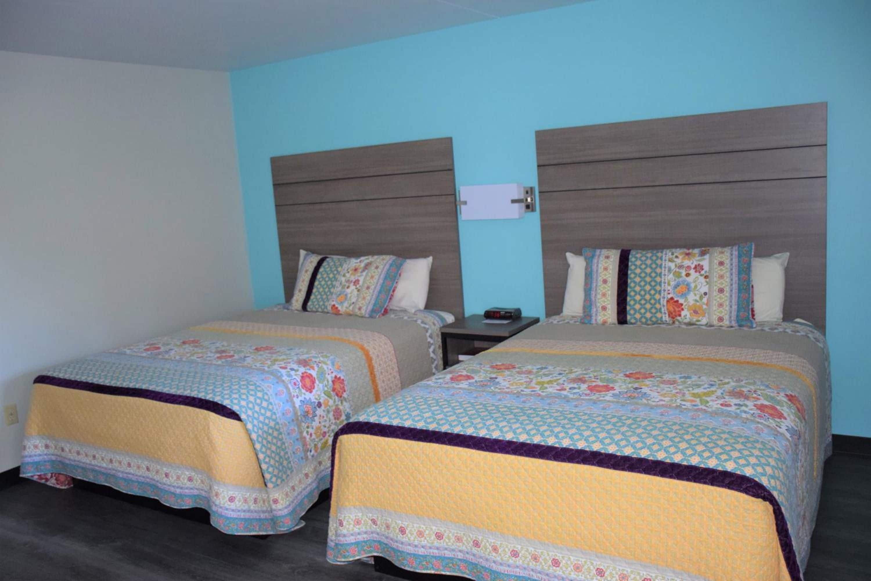 A hotel room with two beds, each with a patterned bedspread and pillows, a nightstand with a lamp, and a wall-mounted power outlet.