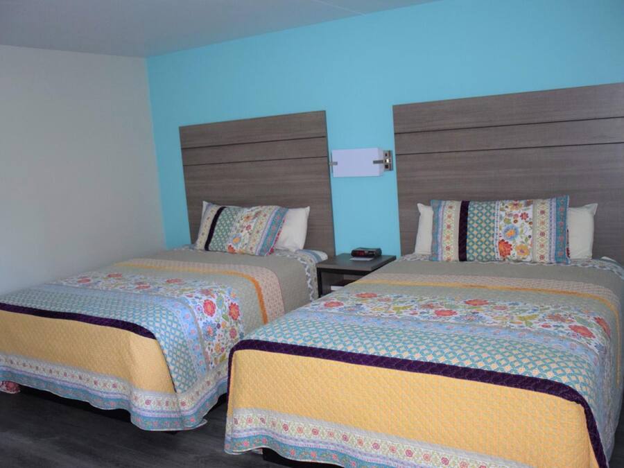 A hotel room with two beds, each with a patterned bedspread and pillows, a nightstand with a lamp, and a wall-mounted power outlet.