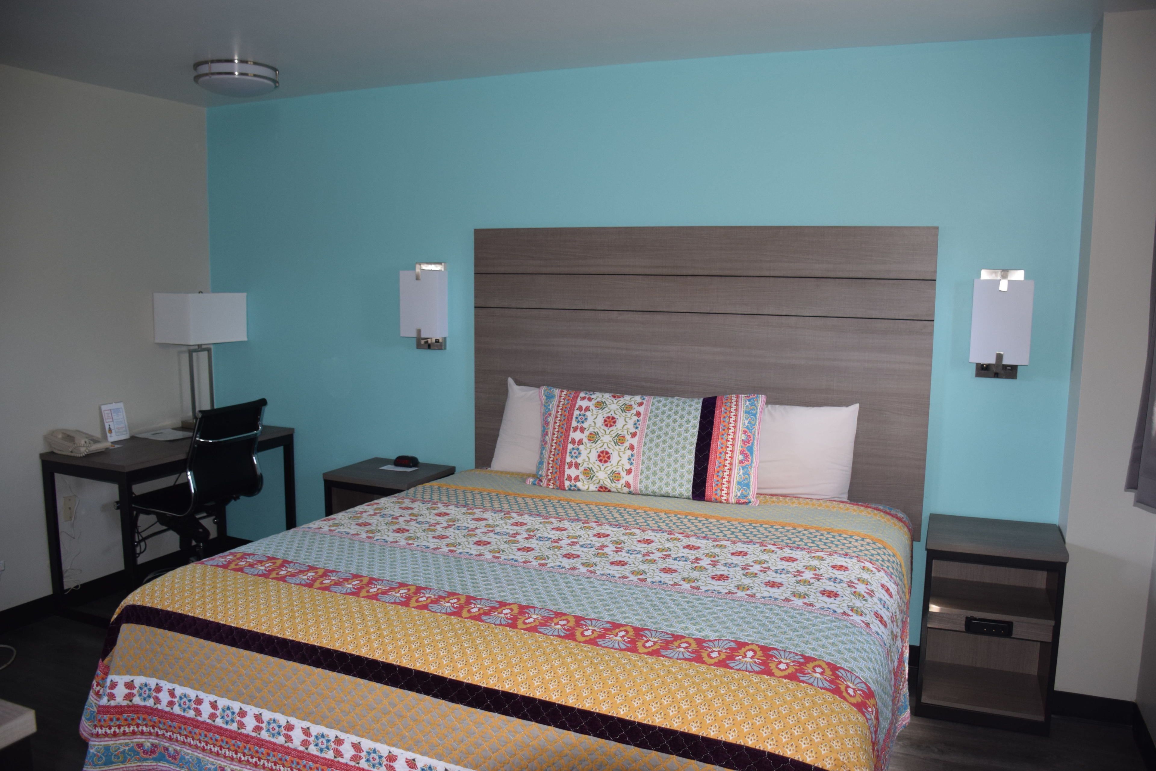 A hotel room with a large bed, a desk with a chair, a nightstand, and a wall-mounted lamp.