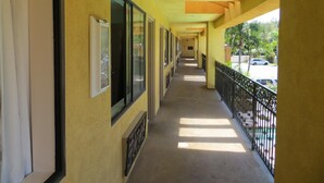 A corridor with large windows, a balcony with a metal railing, and a view of parked cars outside. - Azusa INN Motel (La Puente)