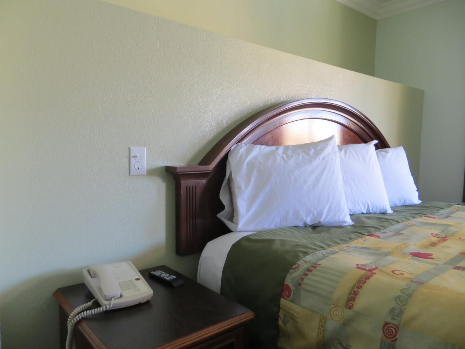 A hotel room with a wooden headboard, a bed with white pillows, a green and yellow patterned bedspread, a bedside table with a telephone and remote control.