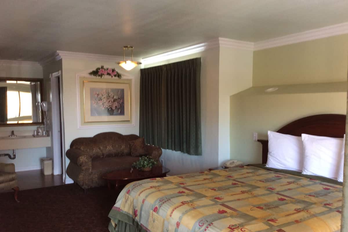 A bedroom with a large bed, a sofa, a small table with a plant, a painting on the wall, and a window with curtains.