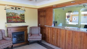 A reception area with a wooden reception desk, two patterned armchairs, a fireplace, and a ceiling fan. - Azusa INN Motel (La Puente)
