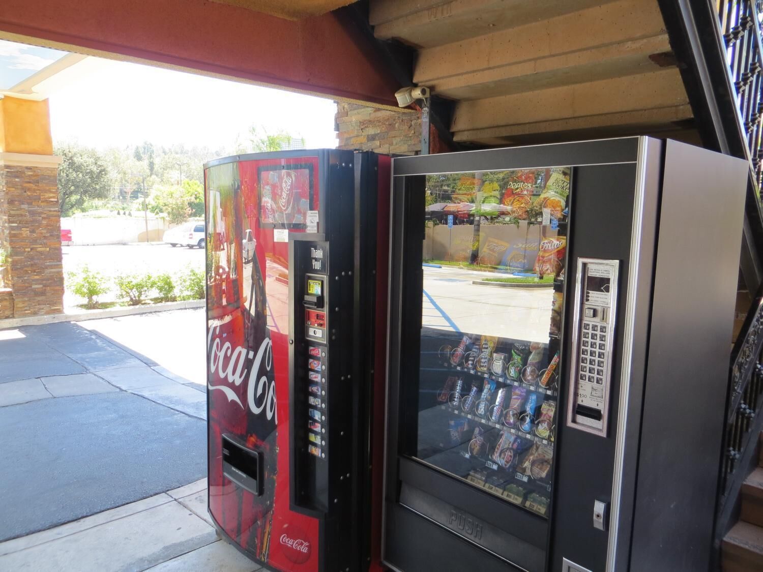 Two vending machines, one Coca-Cola branded and one with a variety of snacks, located outside a building with a brick wall and a covered area.