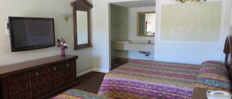 Habitación de hotel con dos camas, un aparador, un televisor, un espejo y un baño a la vista a través de una puerta abierta.