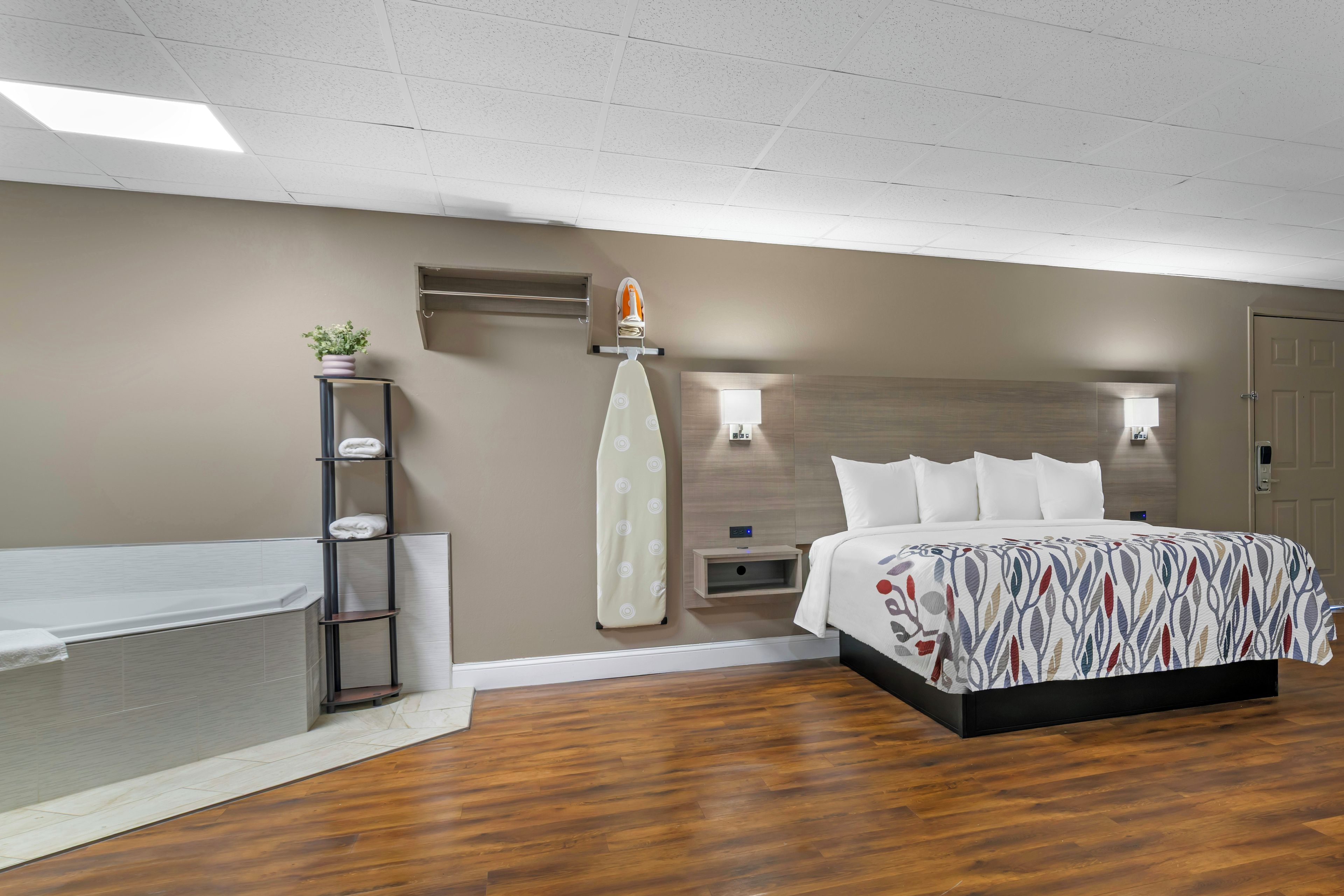 A modern bedroom with a wooden floor, a bed with a patterned bedspread, a nightstand with a lamp, a wall-mounted ironing board, and a small shelf with a plant.