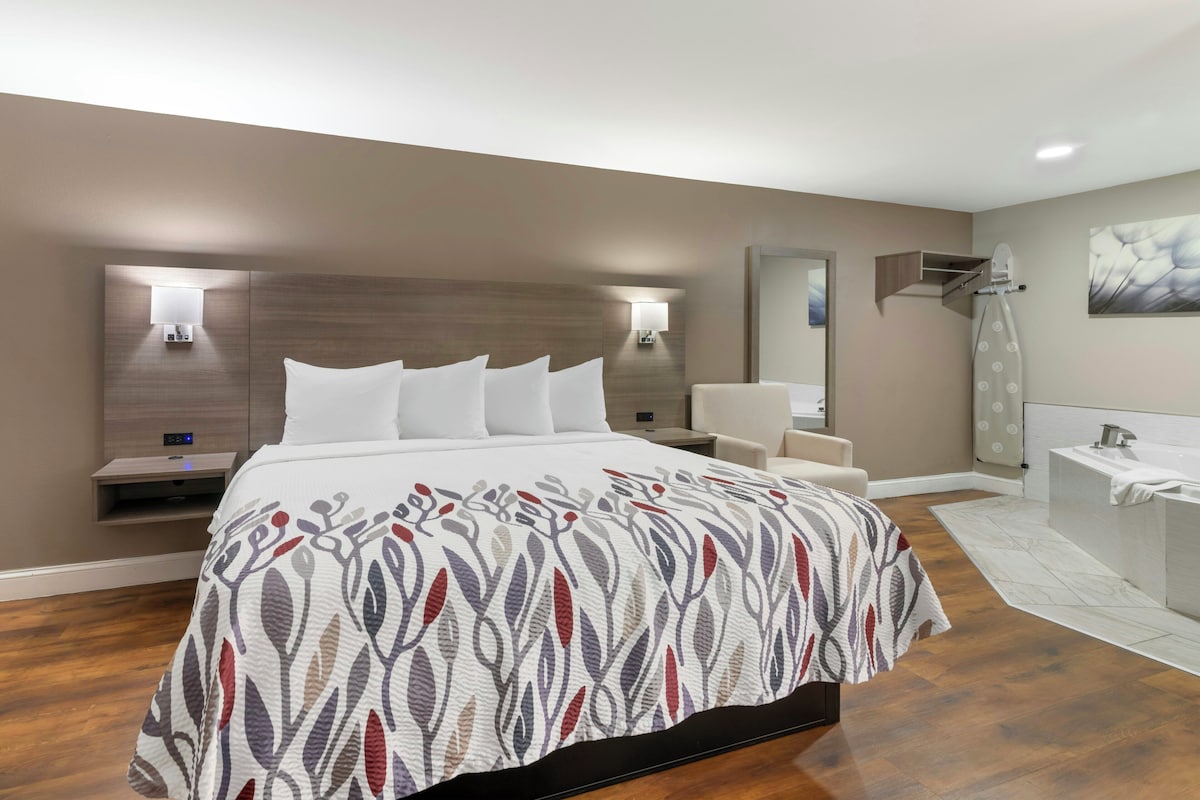 Une chambre d’hôtel moderne dotée d’un grand lit, de tables de chevet, d’une petite table, d’une salle de bain avec douche et d’un luminaire fixé au mur.