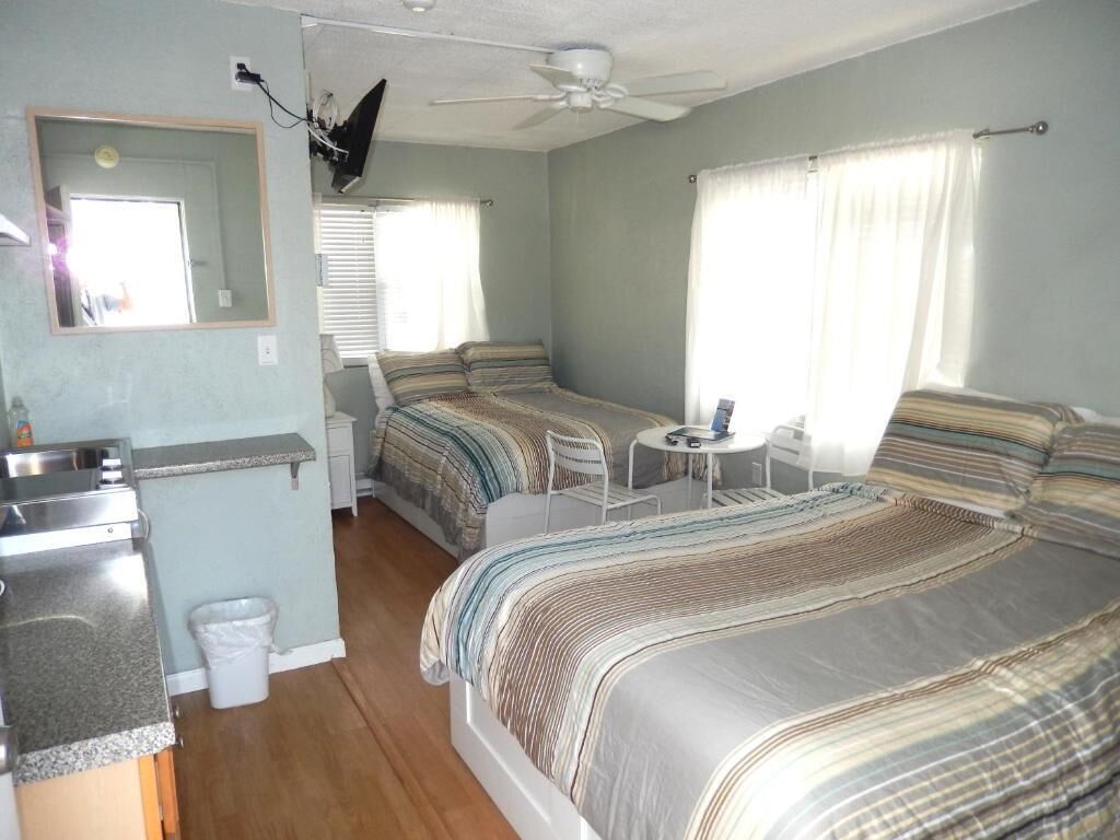 A compact room with a bed, a small table, a chair, a mirror, and a ceiling fan.
