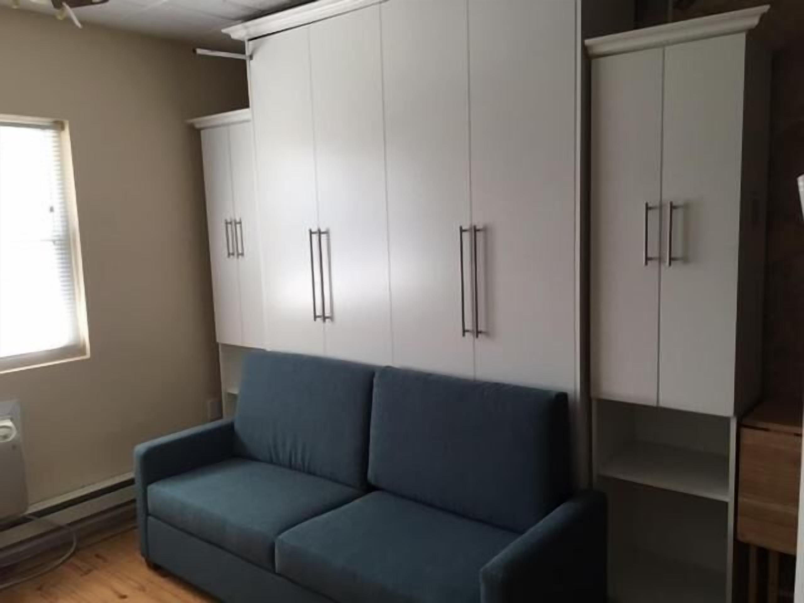 A room with a blue sofa, white cabinets, and a window.