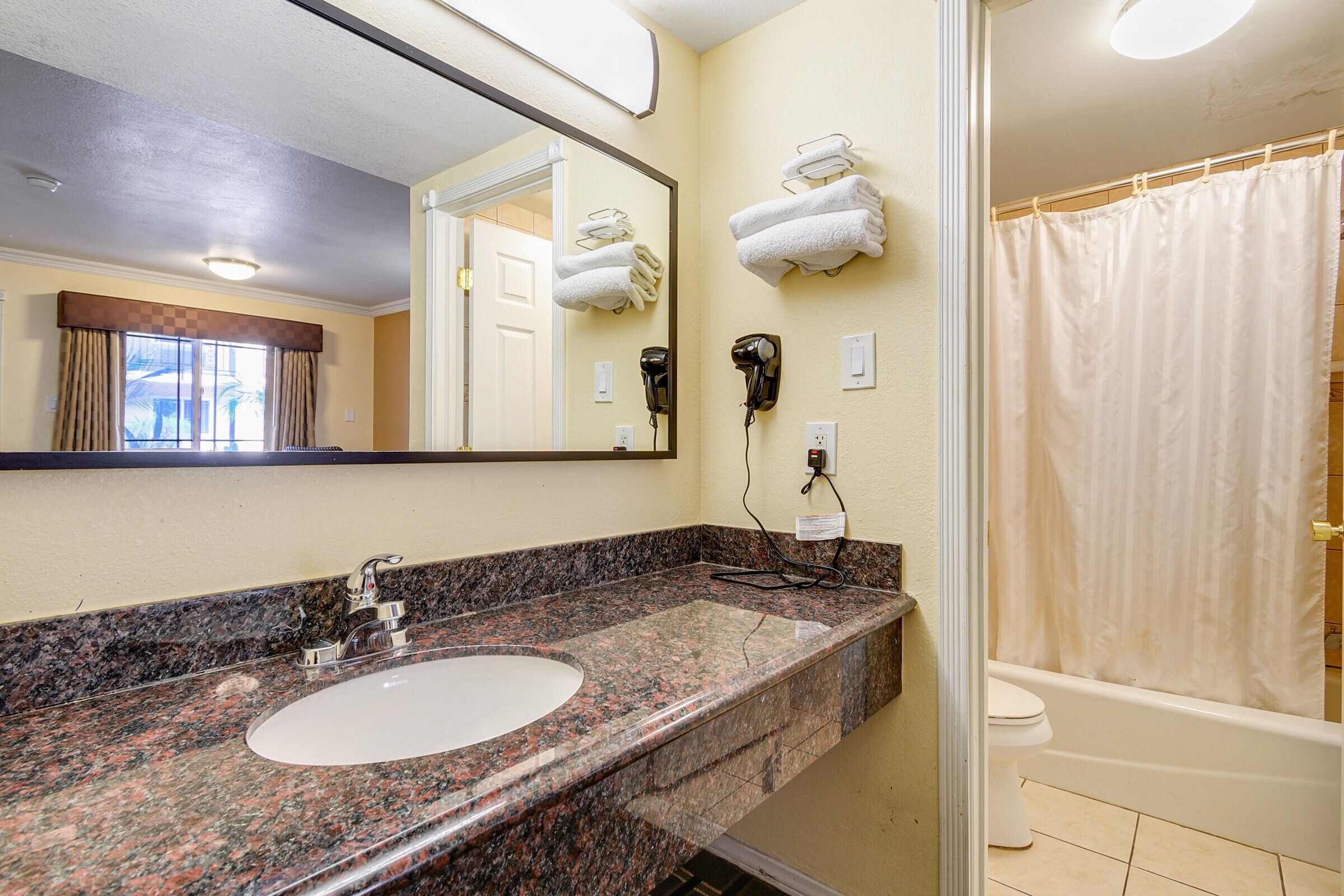 A bathroom with a granite countertop sink, a mirror, a towel rack, a hairdryer, and a shower curtain.