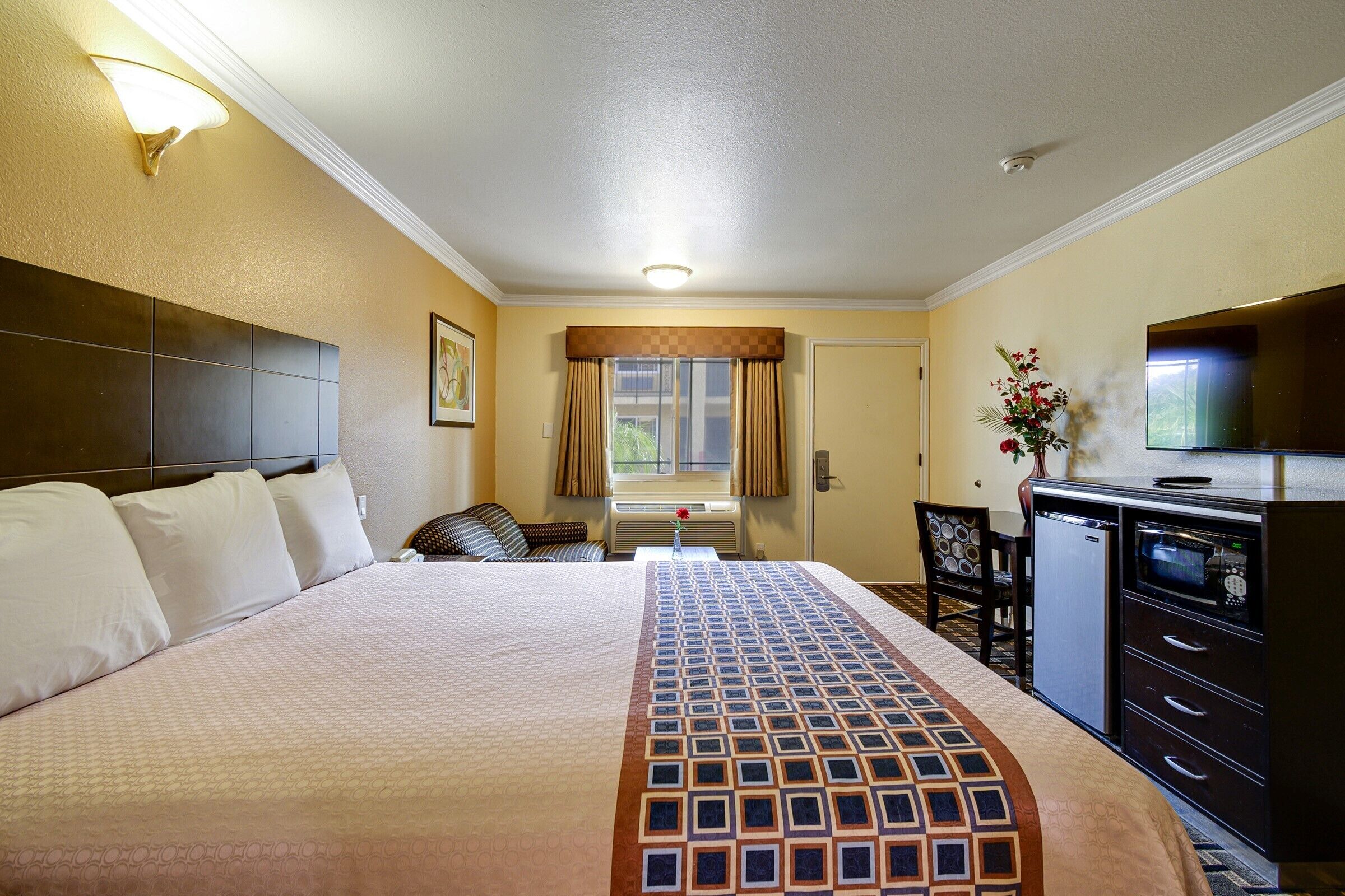 A hotel room with a large bed, a desk with a TV, a small fridge, and a window with curtains.