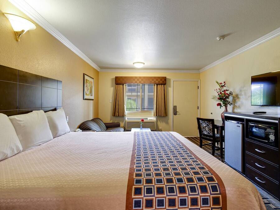 A hotel room with a large bed, a desk with a TV, a small fridge, and a window with curtains.