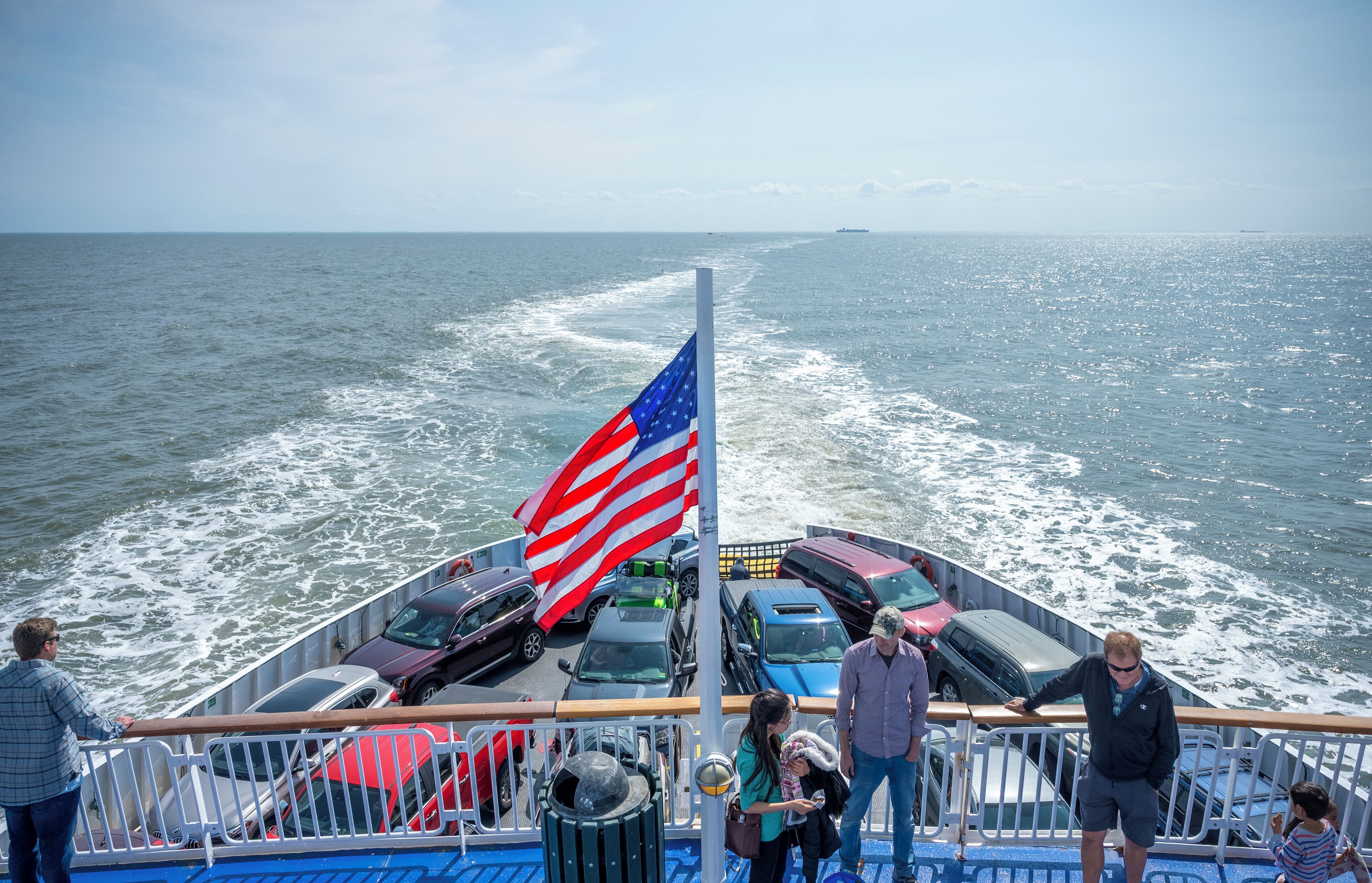 A ferry with multiple cars and passengers, a flag, and a view of the ocean.