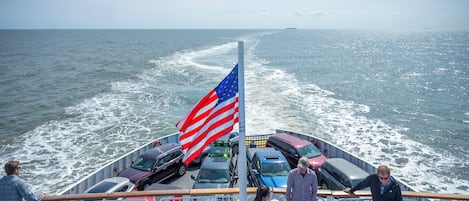 A ferry with multiple cars and passengers, a flag, and a view of the ocean.
