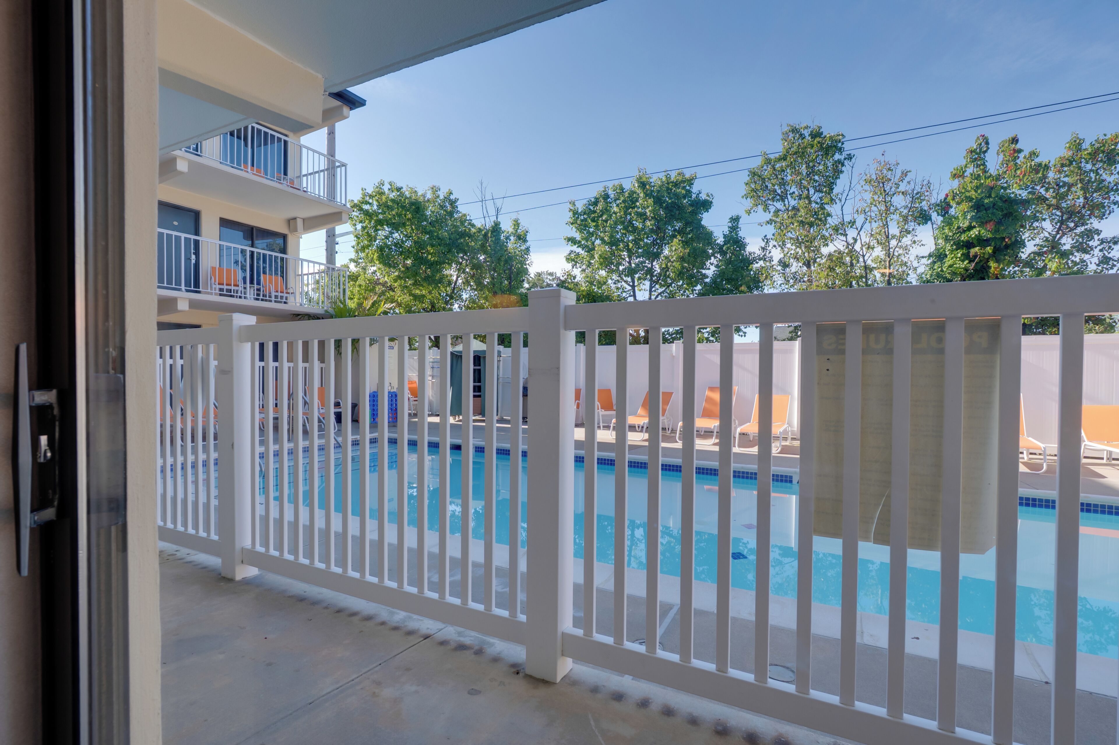 Uma piscina com cerca branca, um edifício com varandas e árvores ao fundo.