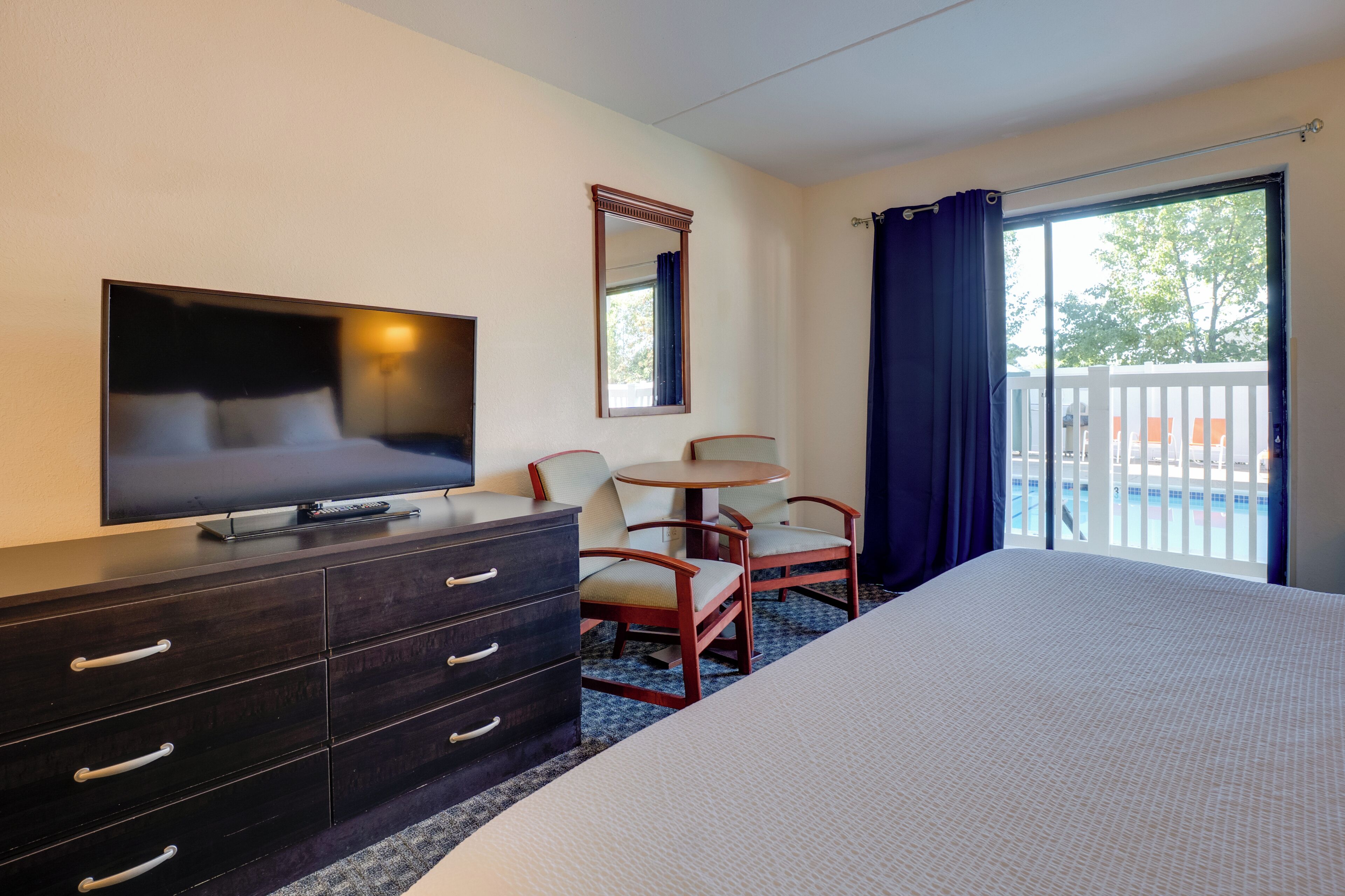 A hotel room with a bed, a TV, a dresser, a chair, a small table, a balcony with a view, and a window with curtains.