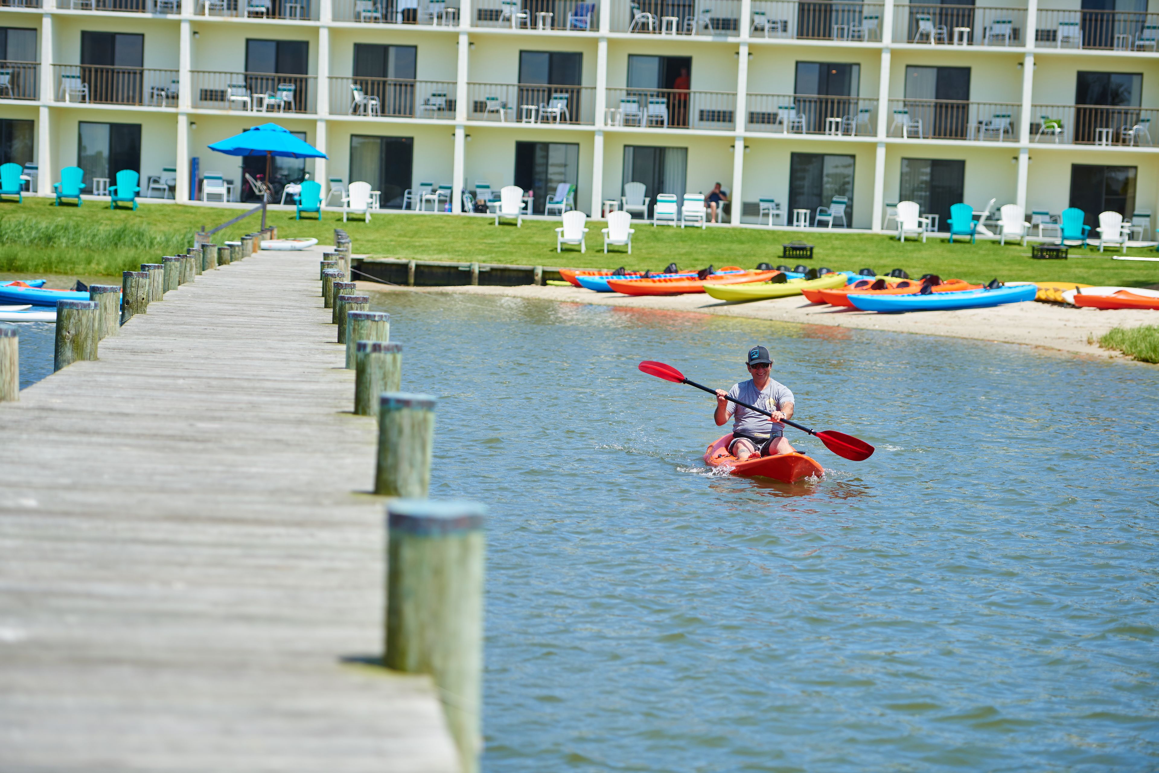 A kayaker on a wooden pier with a row of colorful kayaks lined up on the shore.