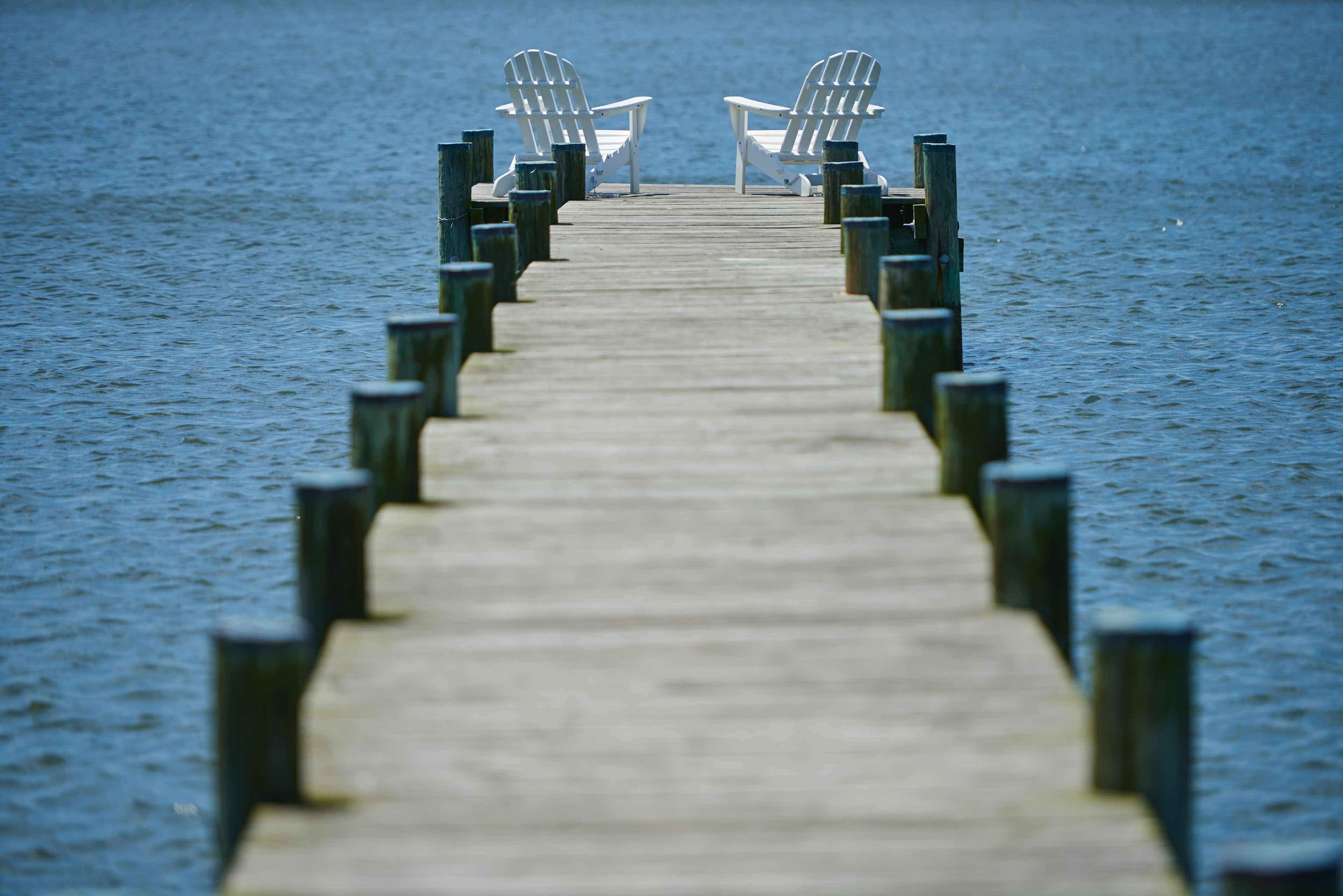 Eine hölzerne Pier, die sich über ein Gewässer erstreckt, mit zwei weißen Stühlen am Ende.
