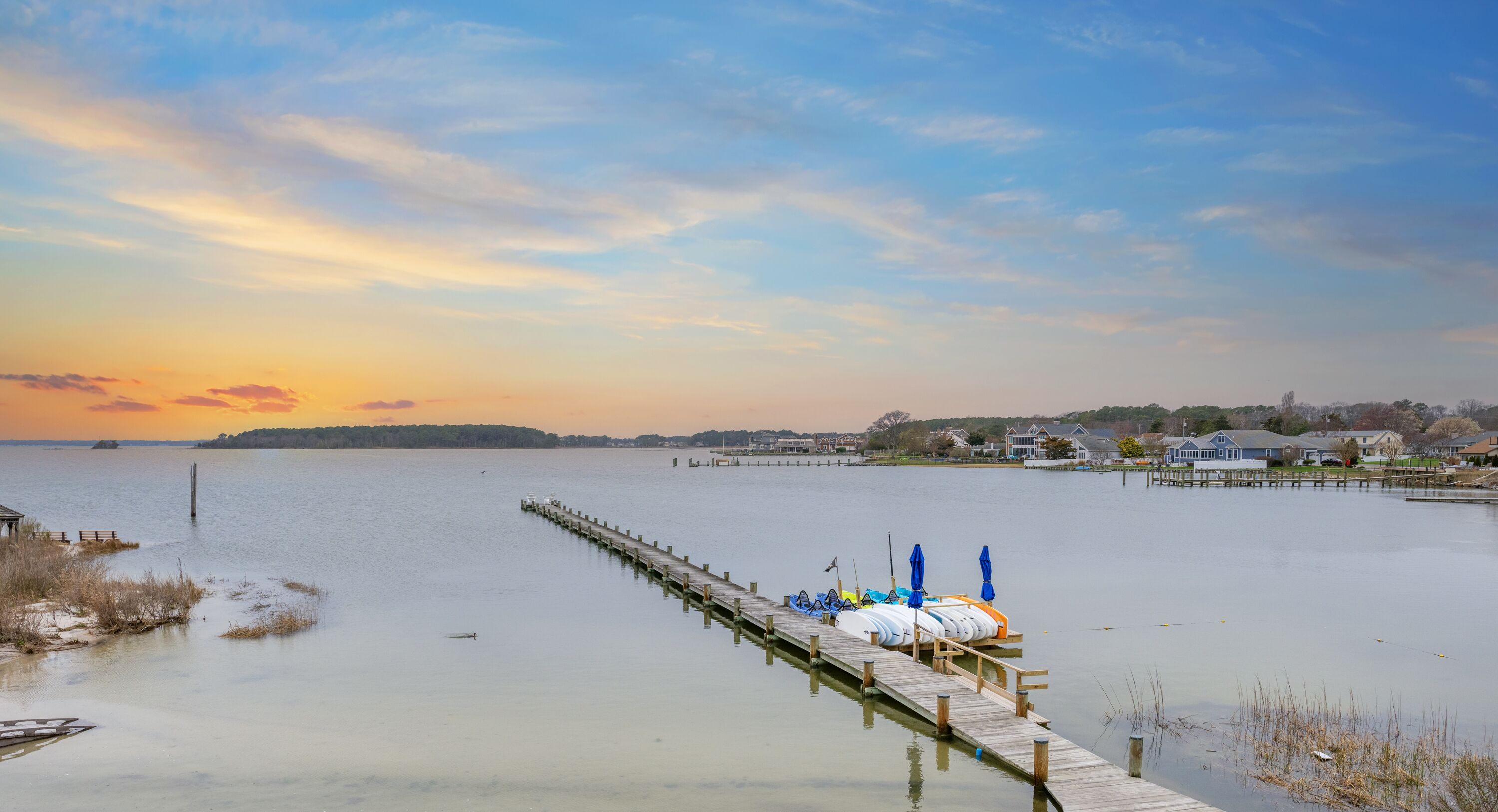 A wooden pier extending into calm water with boats docked at the end, surrounded by a residential area with houses and a clear sky.