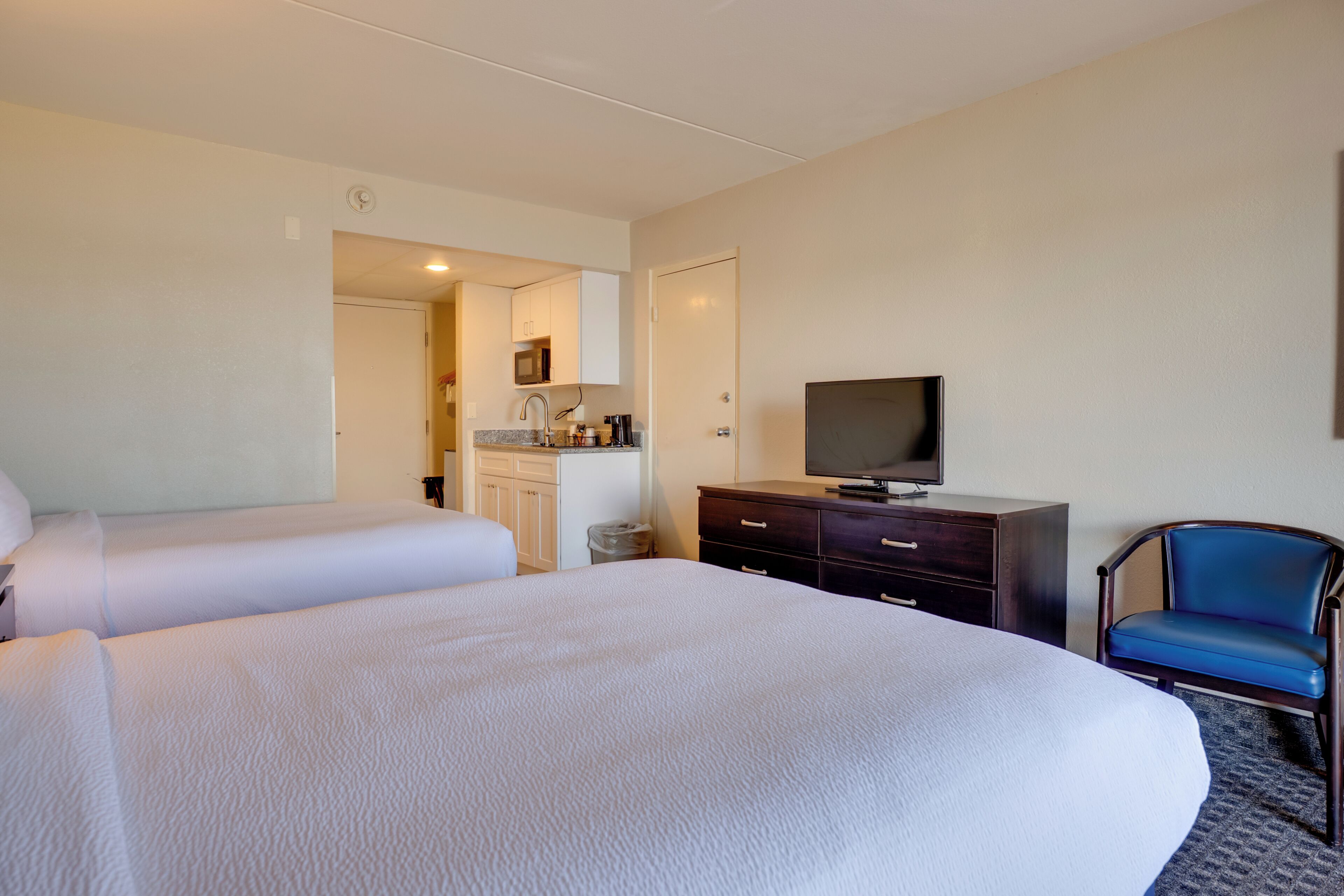 A hotel room with two beds, a television, a desk with a chair, and a kitchenette.