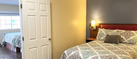 A hotel room with a bed, a nightstand, a wall-mounted lamp, and a door leading to another room.
