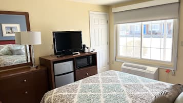 Habitación de hotel con cama, televisor sobre un soporte de madera, escritorio con lámpara, mini refrigerador, ventana con vistas a edificios y una puerta.